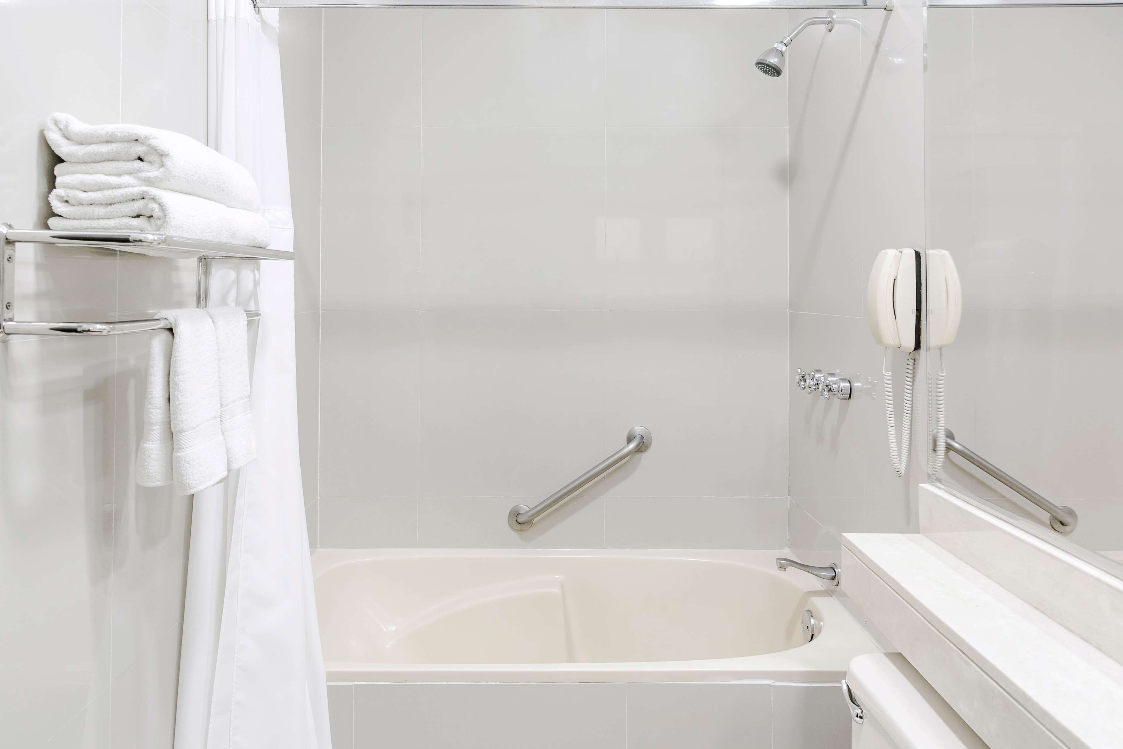 Une salle de bain moderne avec une baignoire, une douche et des toilettes. Des serviettes blanches sont pliées et empilées sur une étagère, et un téléphone est fixé au mur.
