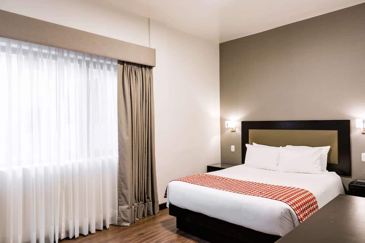 Ein Hotelzimmer mit einem großen Bett, einem Nachttisch, einer Wandleuchte und einem Fenster mit Vorhängen.