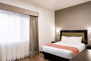Una camera d'albergo con un letto grande, un comodino, una lampada a parete e una finestra con tende.