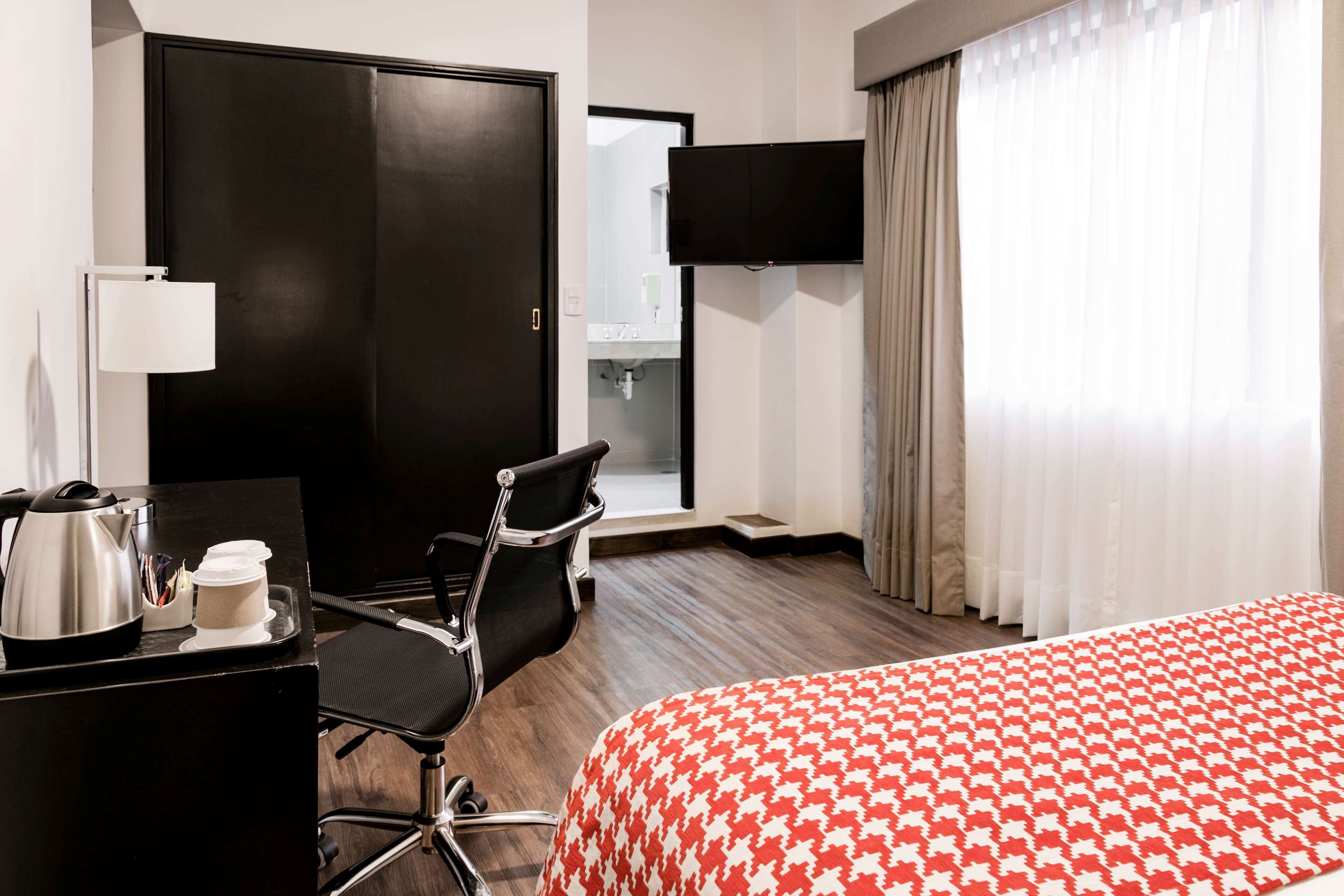 Habitación de hotel con cama, escritorio con hervidor y tazas, silla, televisor y baño visible a través de una puerta abierta.