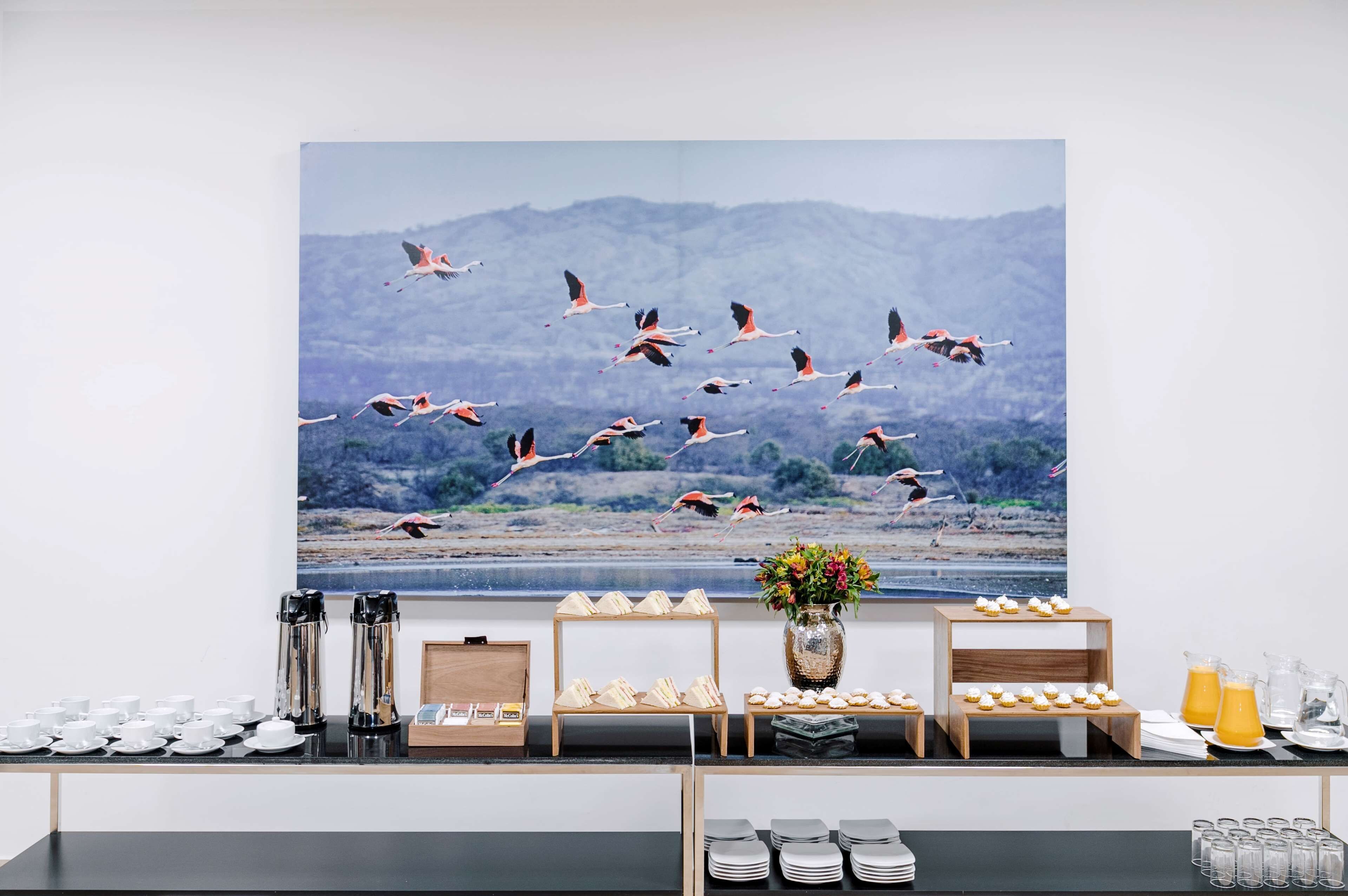A buffet setup with various food items and beverages, a large painting of birds in flight, and a vase of flowers.