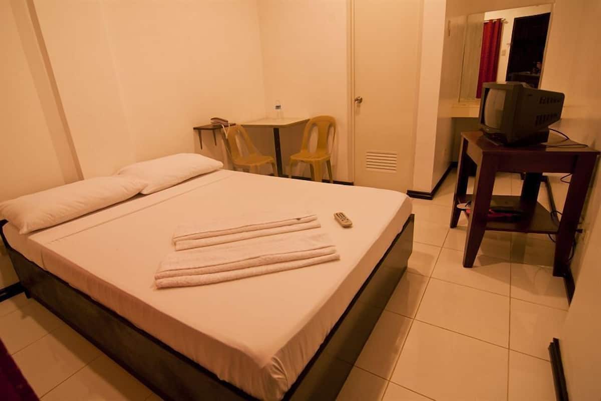 A single bed with a white mattress and pillows, a small table with a chair, a television on a wooden stand, and a mirror on the wall.