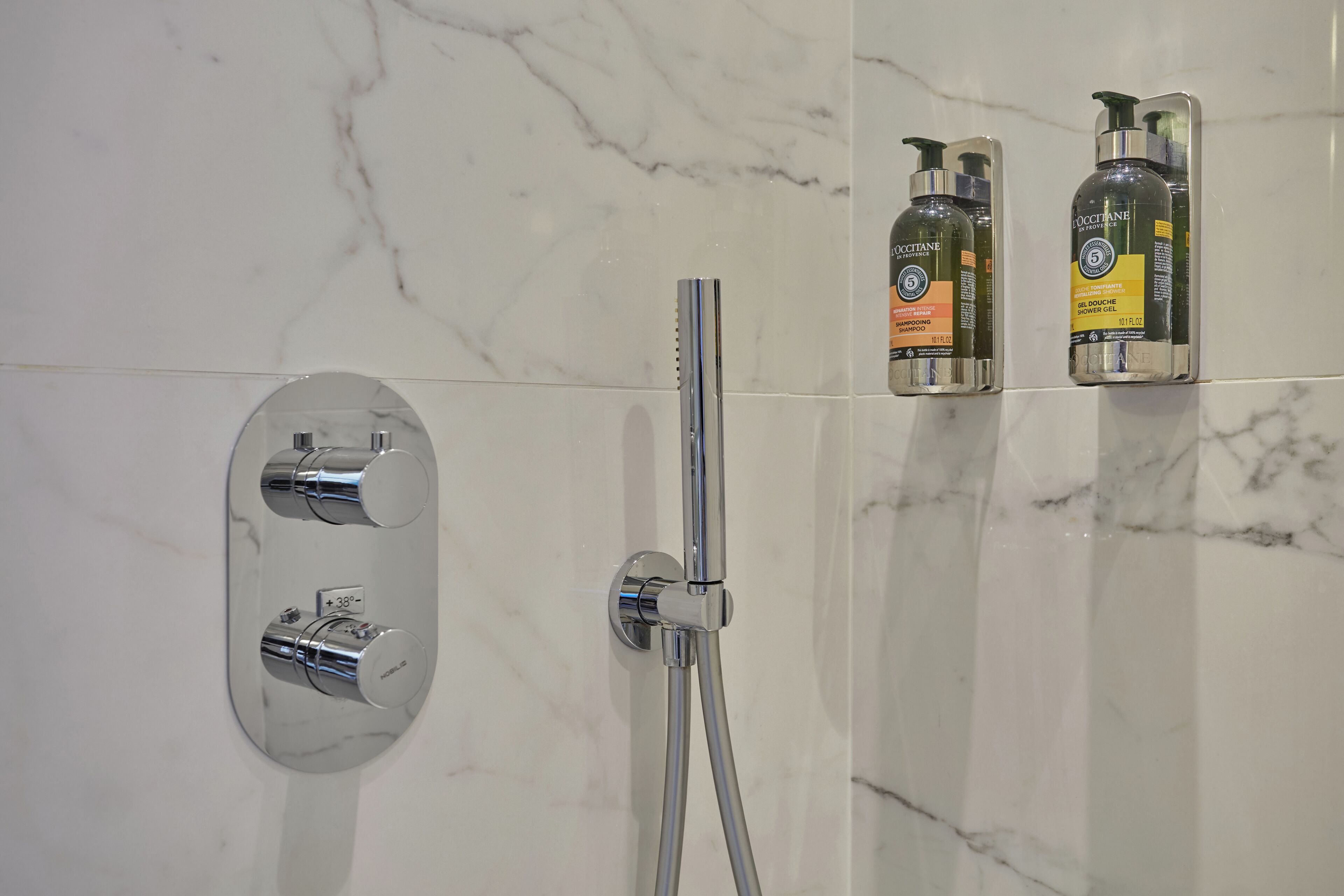 A bathroom with a marble wall, a shower with a handheld showerhead, and two bottles of hair care products.