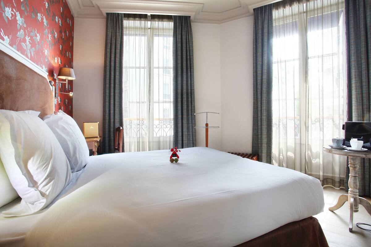 Una cama bien hecha con sábanas blancas, una pequeña figura roja de juguete sobre la cama, y una mesita con una tetera y una taza a la derecha.