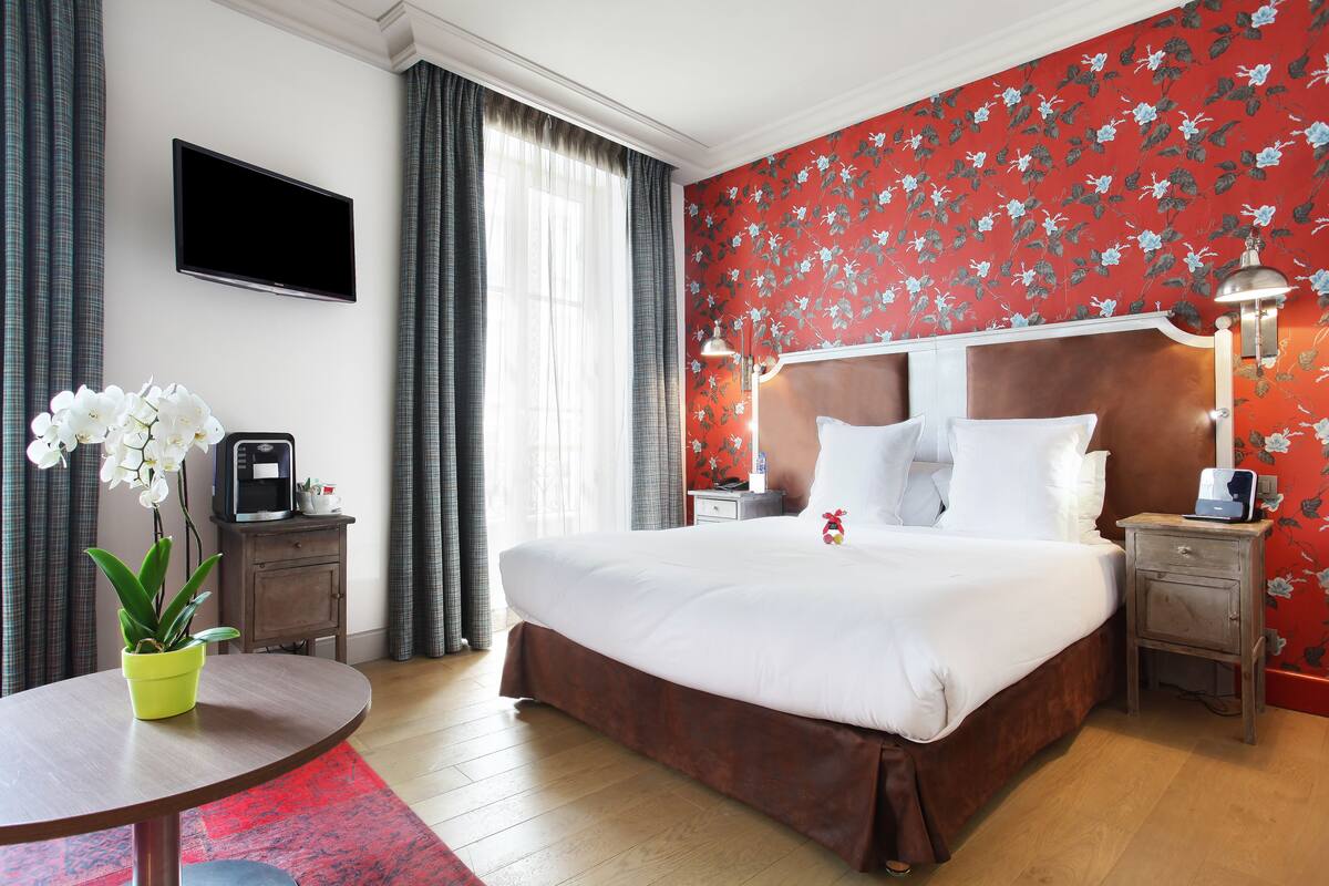 Habitación de hotel con una cama grande, un televisor de pantalla plana montado en la pared, una mesita de noche con lámpara y una pequeña mesa con una planta en maceta.