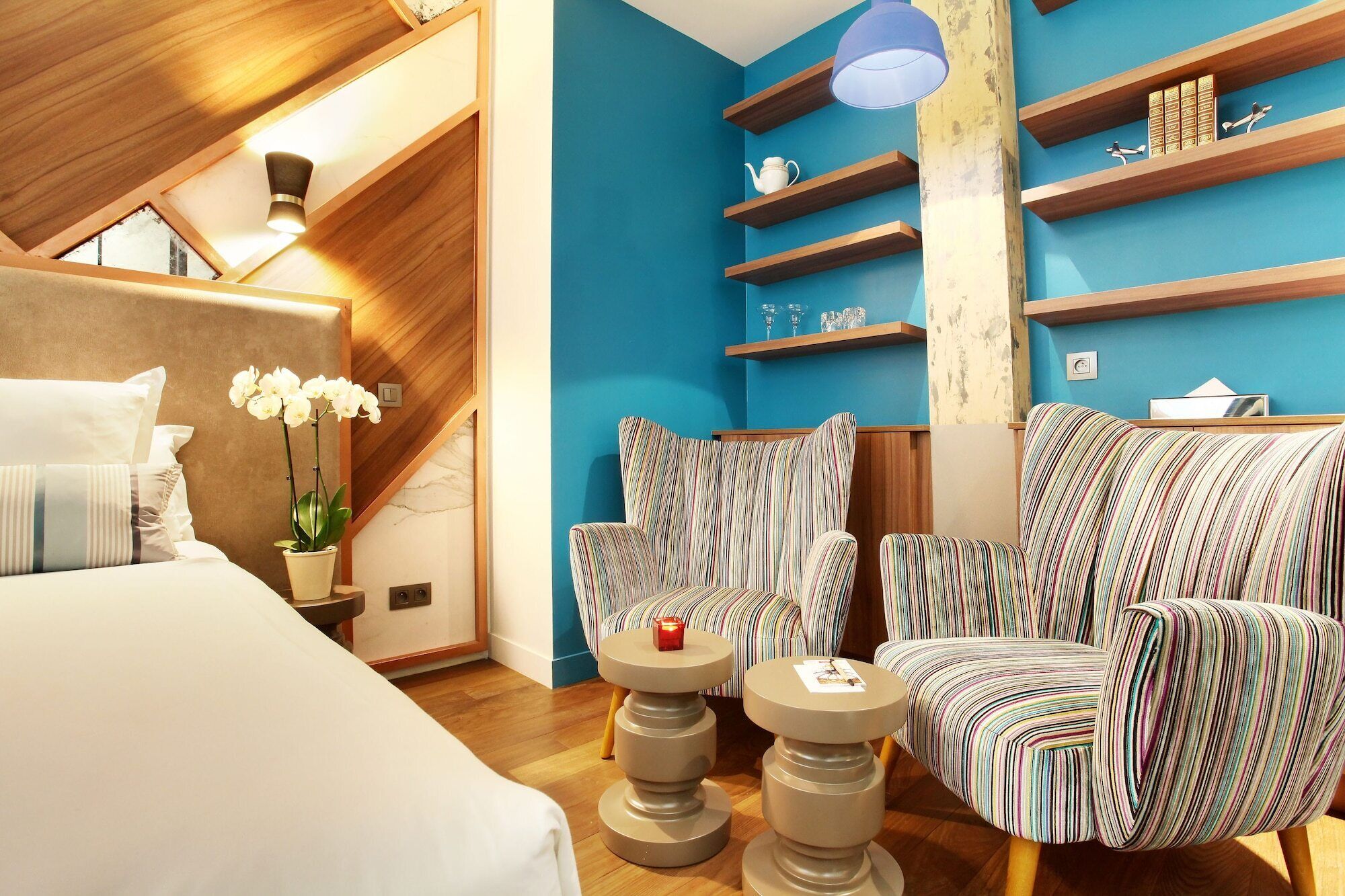 A modern hotel room with a wooden ceiling, a blue accent wall, a bed with white linens, striped armchairs, and a small table with a candle.