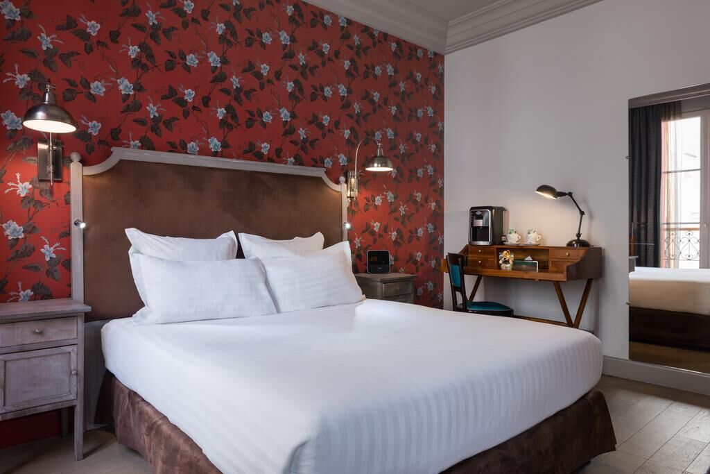 Een net opgemaakt bed met witte lakens, een houten hoofdbord en een behang met bloemen. Er is een nachtkastje met een lamp, een bureau met een lamp, en een raam met gordijnen.