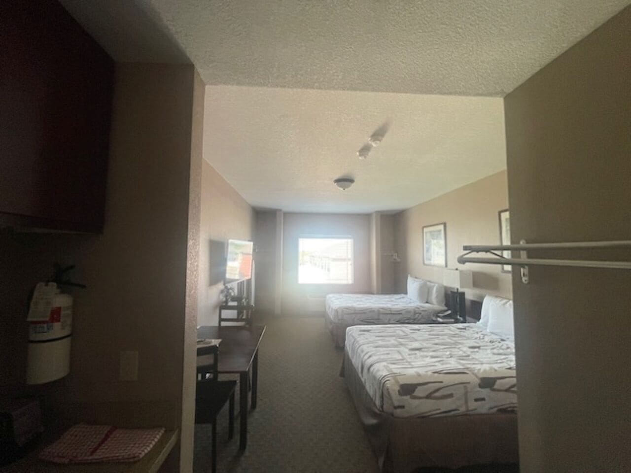 A hotel room with two beds, a desk, a chair, and a window providing natural light.
