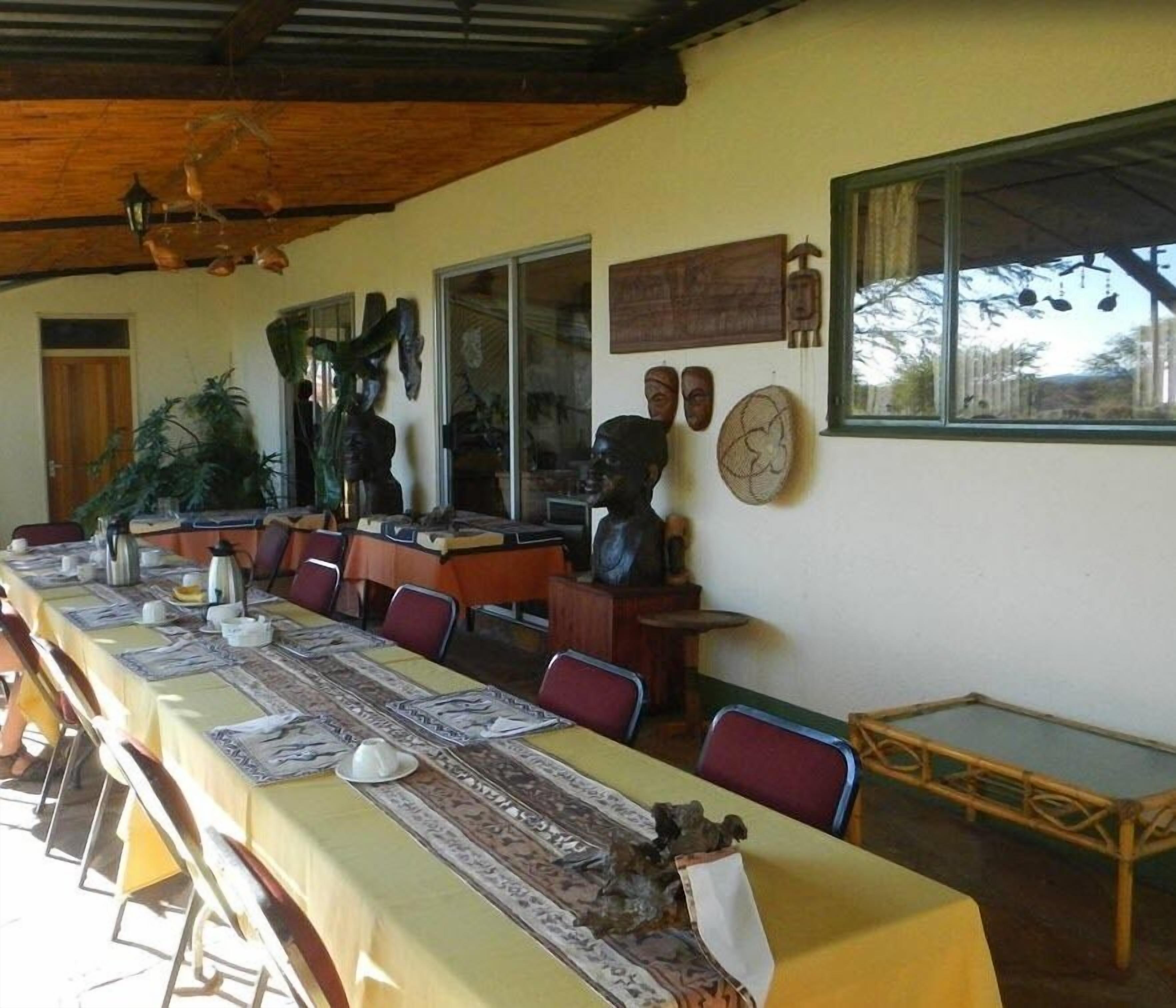 A dining area with a long table covered with a yellow tablecloth, surrounded by chairs. There are decorative items on the walls, including masks and a clock.