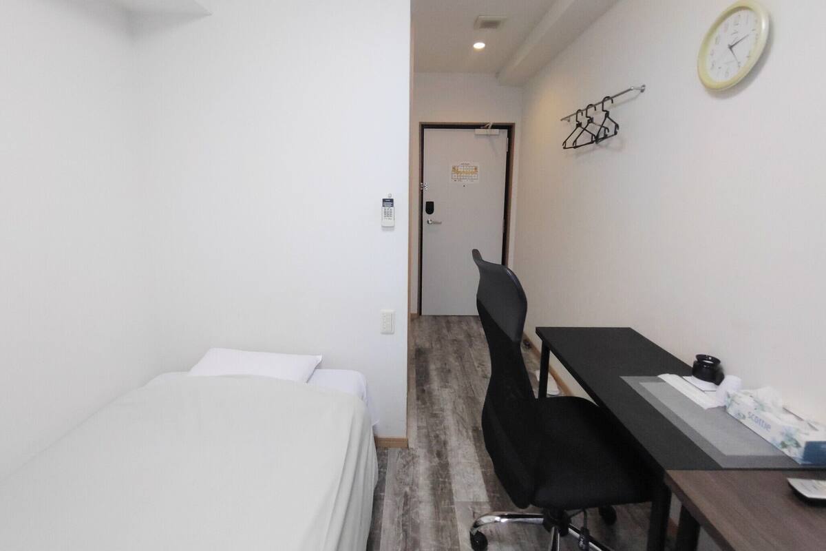 A small, clean room with a bed, a desk, a chair, a clock, and a door.