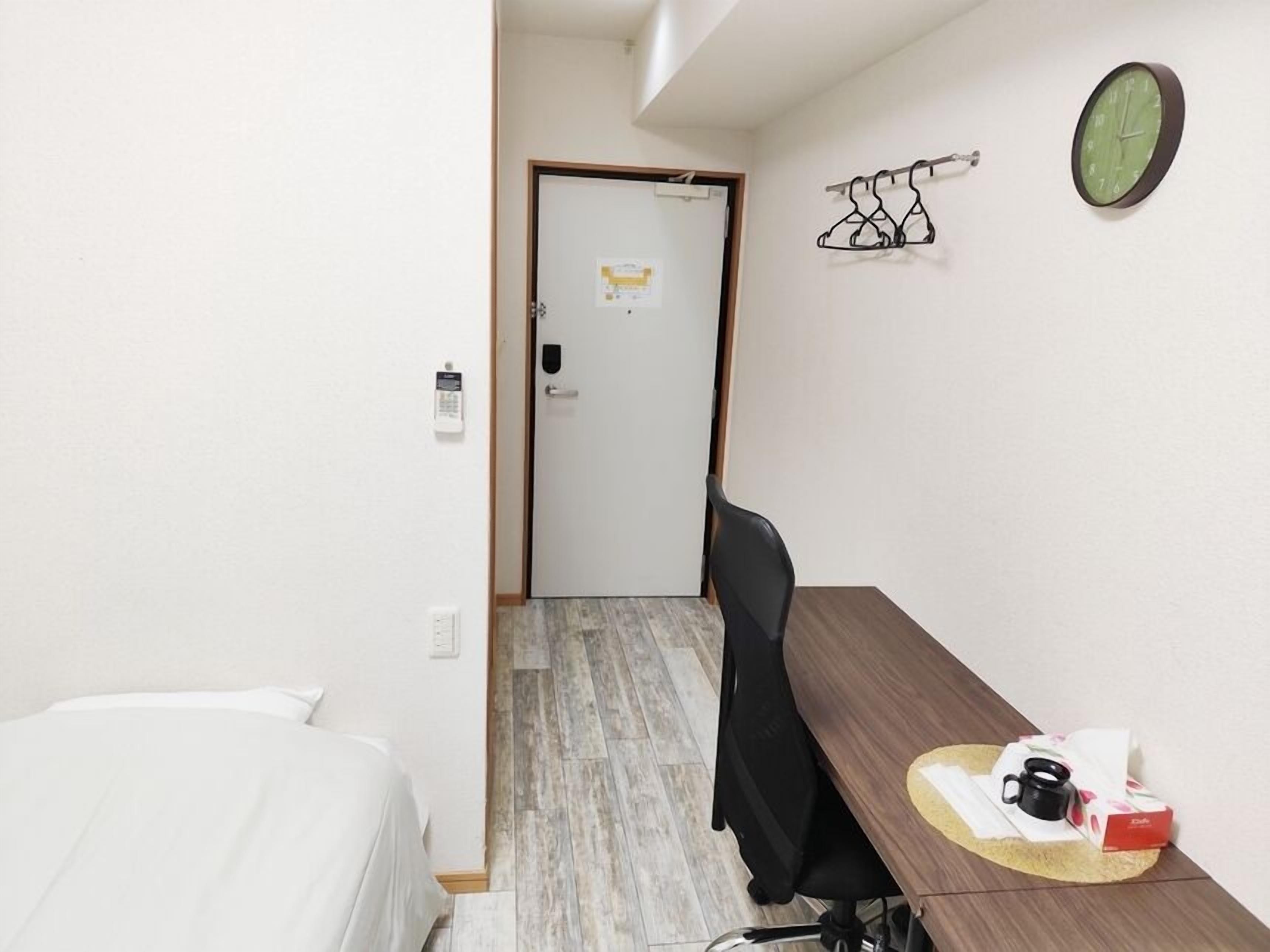 A hotel room with a bed, a desk with a chair, a clock, and a coat rack.