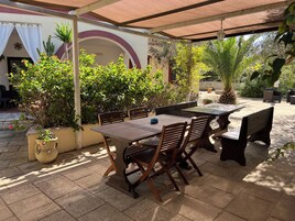 Un patio al aire libre con mesa y sillas de madera, rodeado de plantas en macetas y una pérgola.