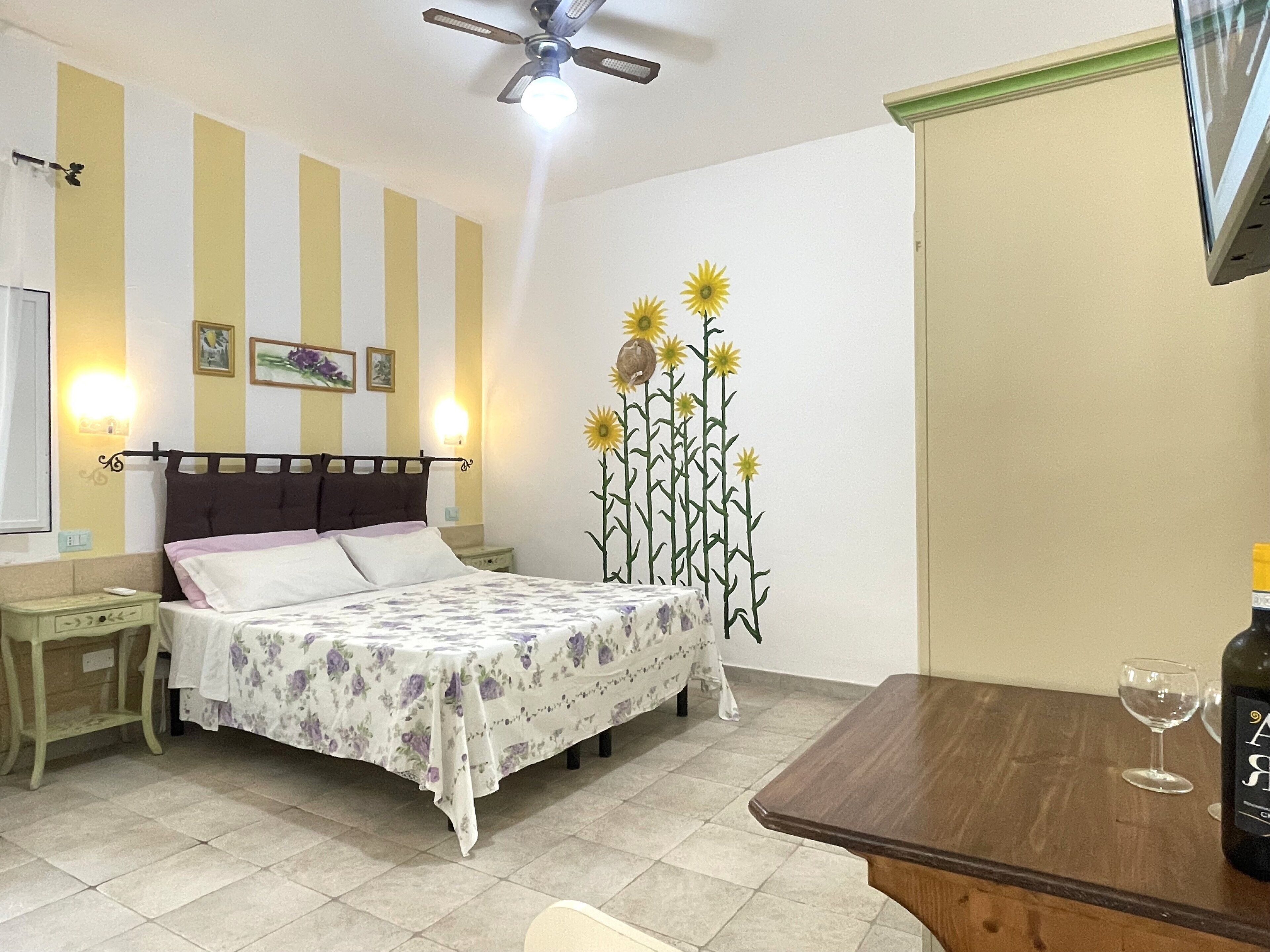 Un dormitorio con cama, mesitas de noche, televisor montado en la pared, ventilador de techo y un mural decorativo con girasoles.