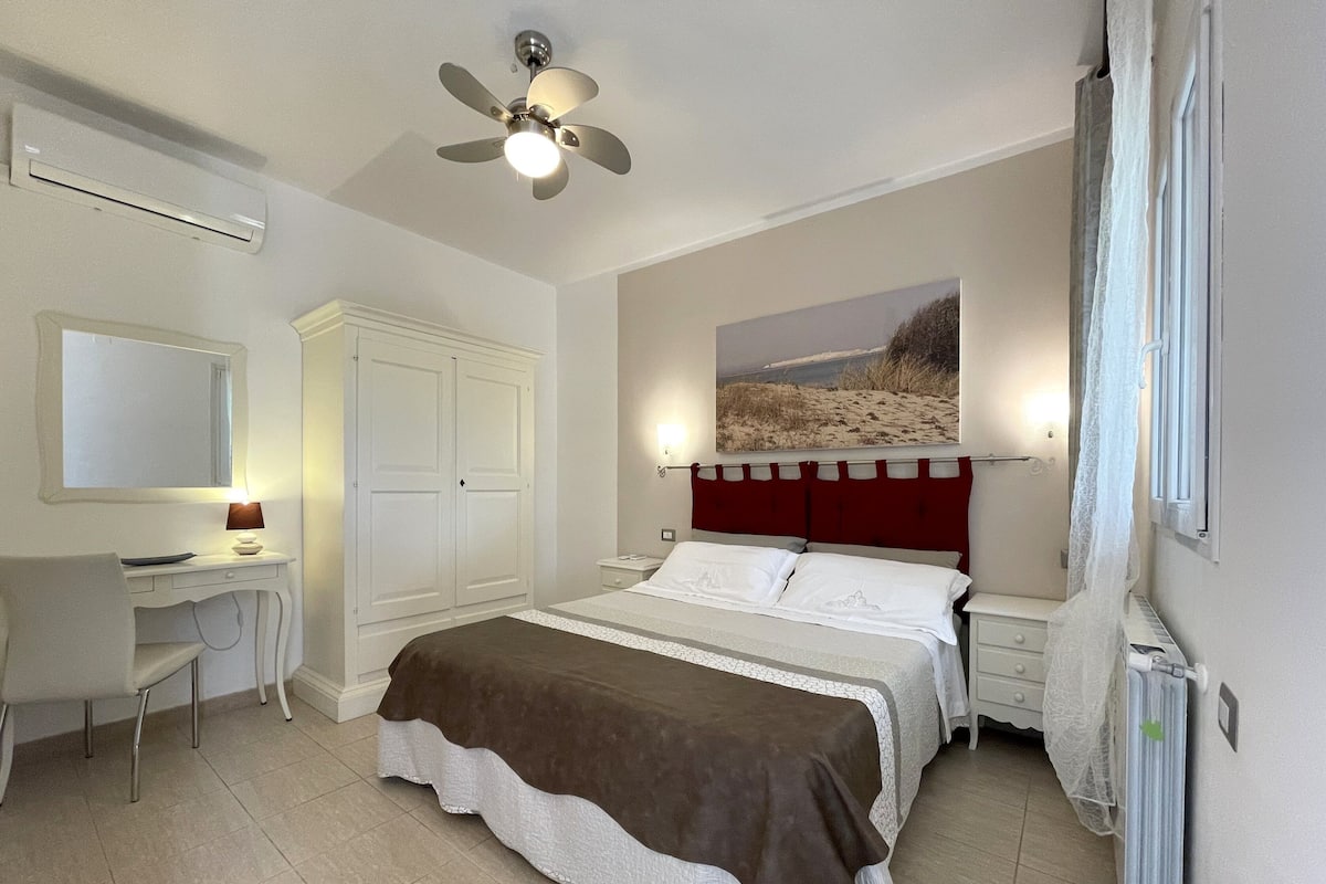 A bedroom with a bed, a desk, a chair, a wardrobe, a ceiling fan, a radiator, and a painting on the wall.