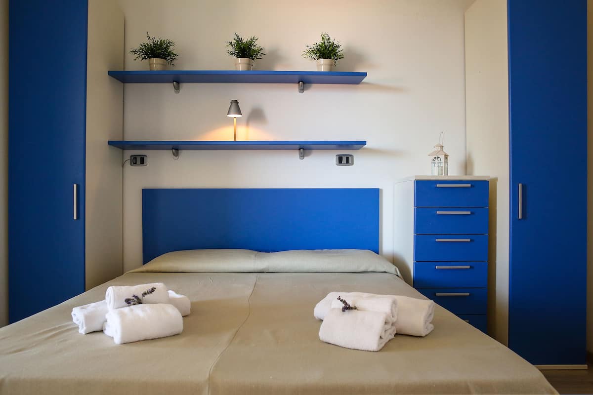 A bedroom with a bed, two blue nightstands, a dresser, and wall-mounted shelves with plants.
