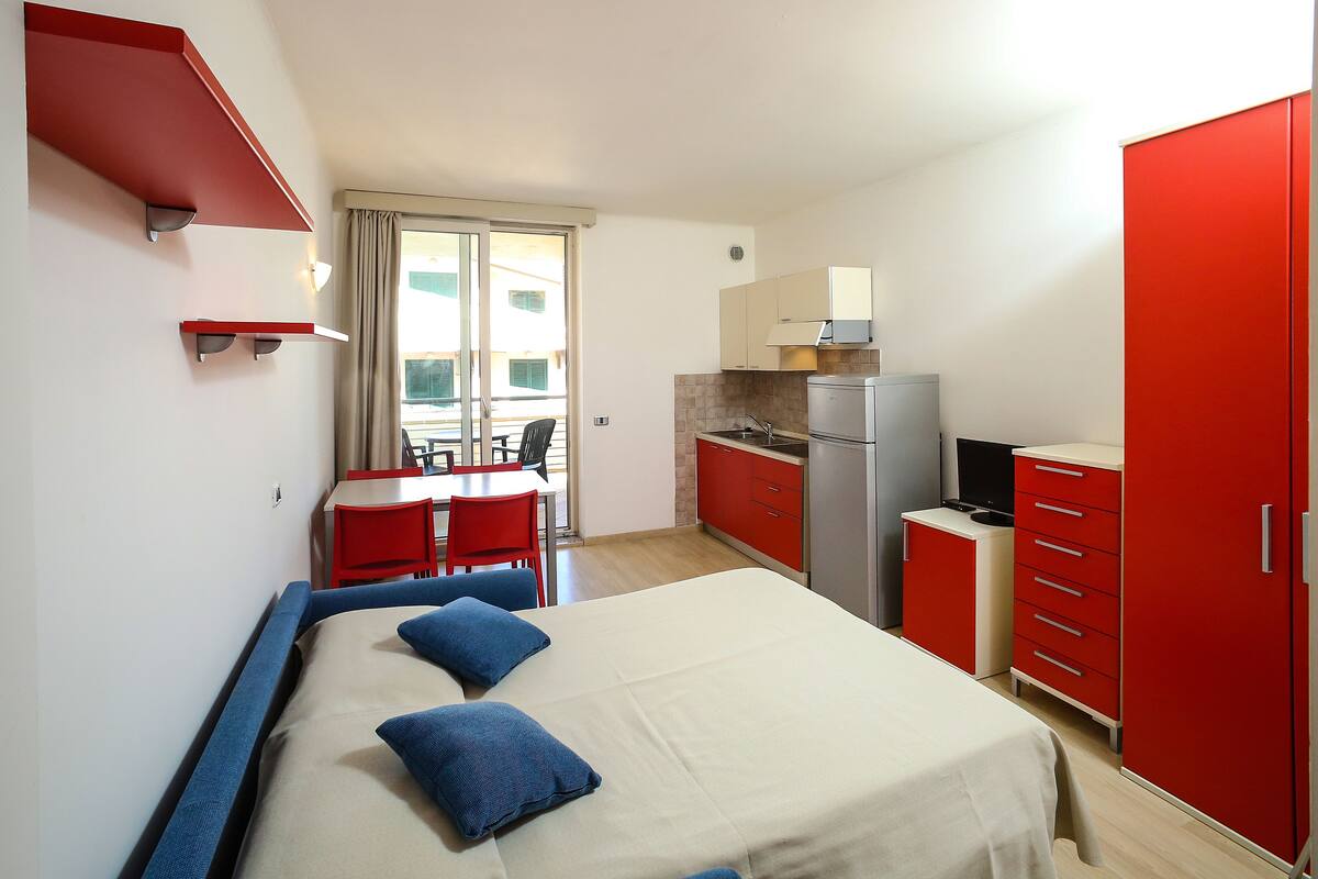 A compact hotel room with a bed, a desk, a chair, a kitchenette, and a wardrobe.