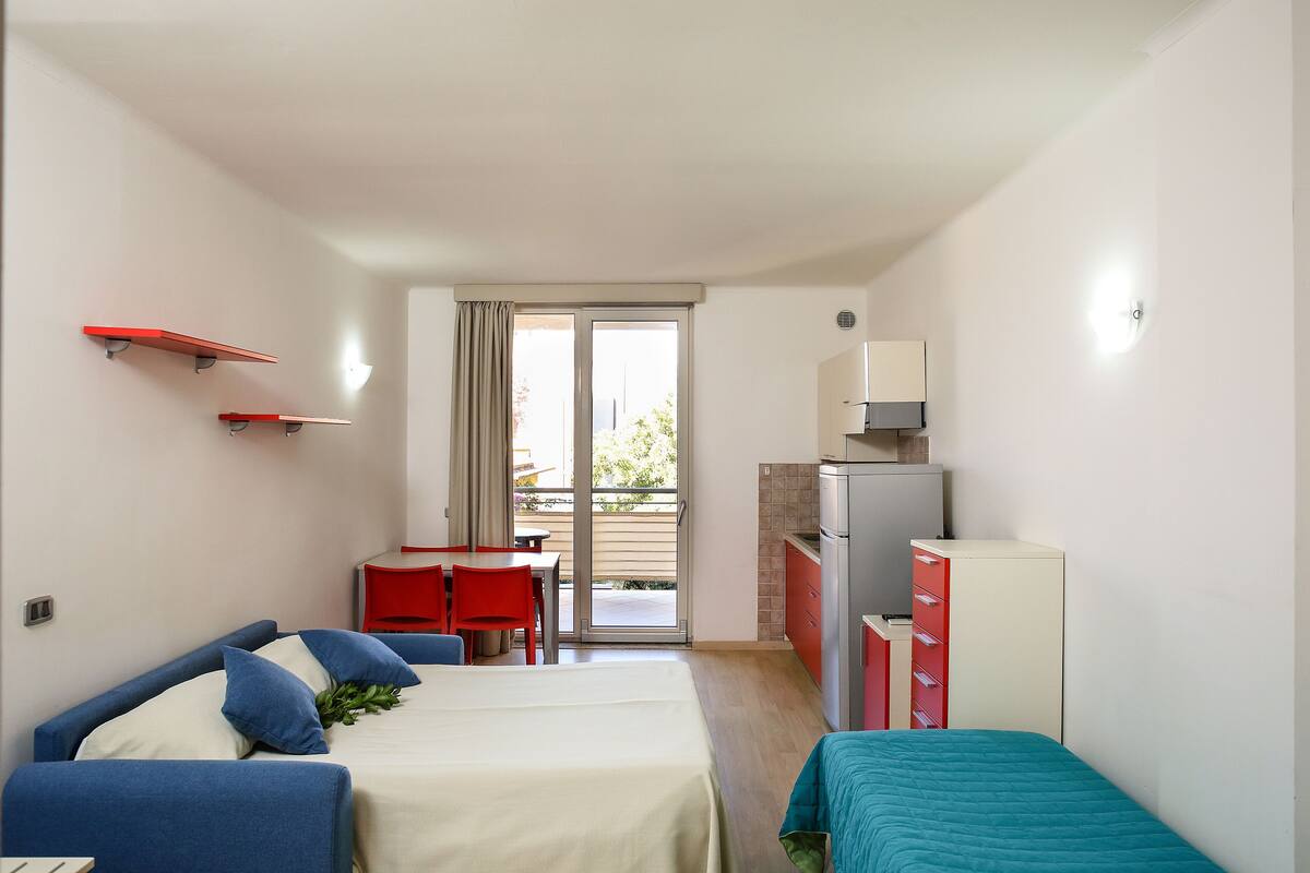 A hotel room with two beds, a desk, a kitchenette, and a sliding glass door to the balcony.