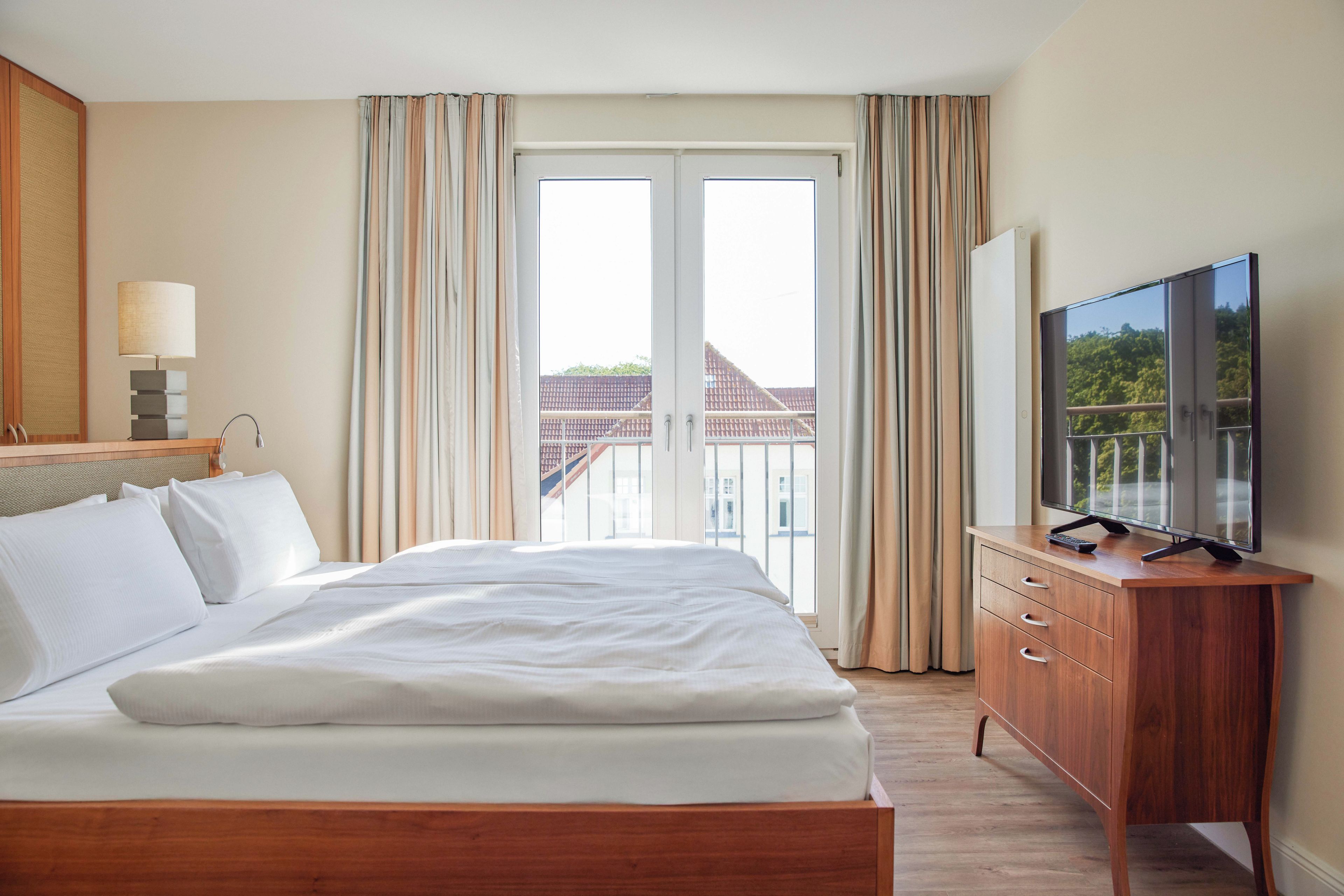 Ett hotellrum med en säng, en tv-apparat på en byrå i trä och utsikt över byggnader genom fönstret.