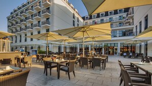 Een terras met tafels en stoelen, parasols en een gebouw met balkons op de achtergrond.
