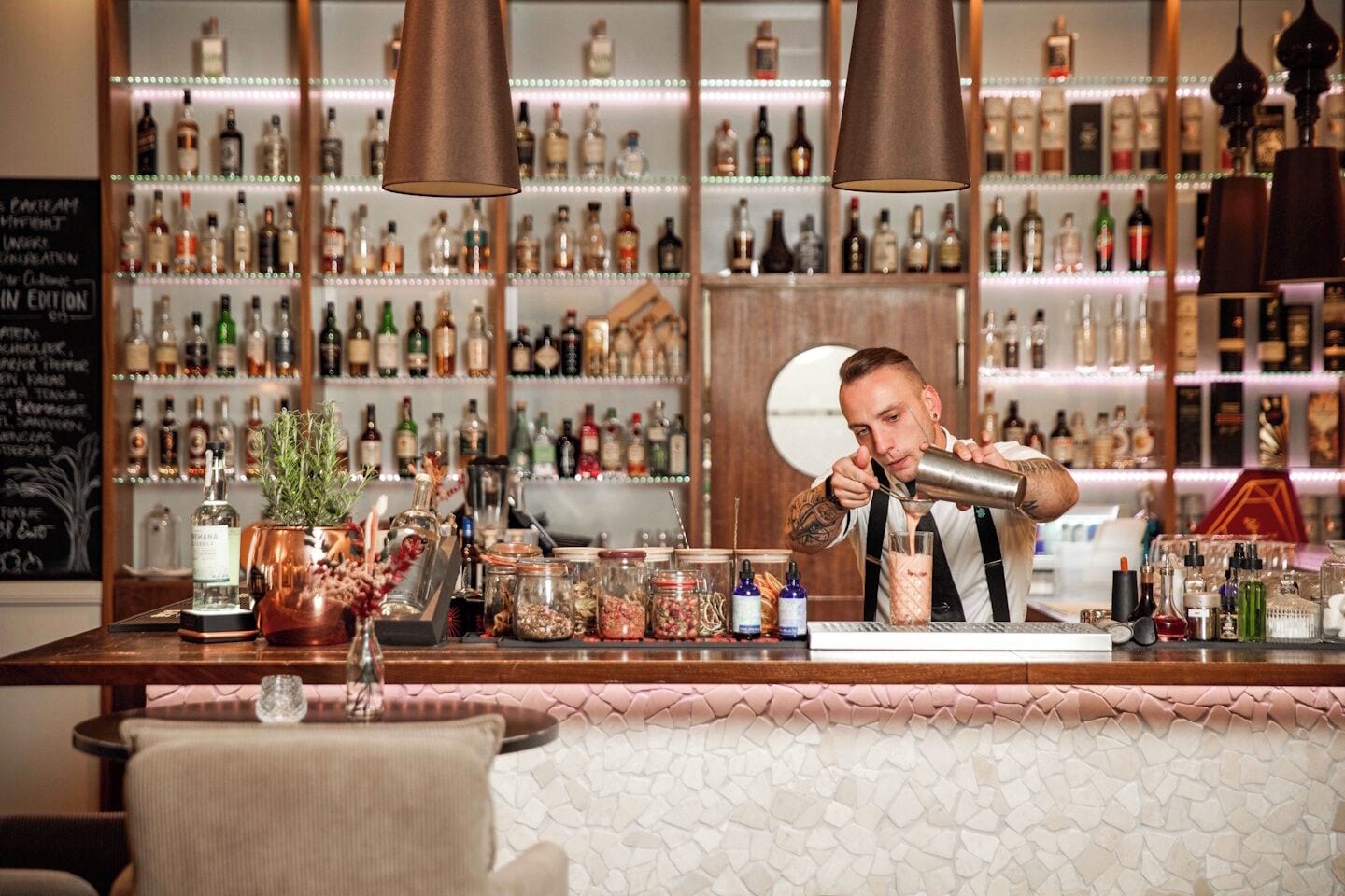 A bar area with a bartender preparing a drink, shelves of liquor bottles, and a chalkboard menu.