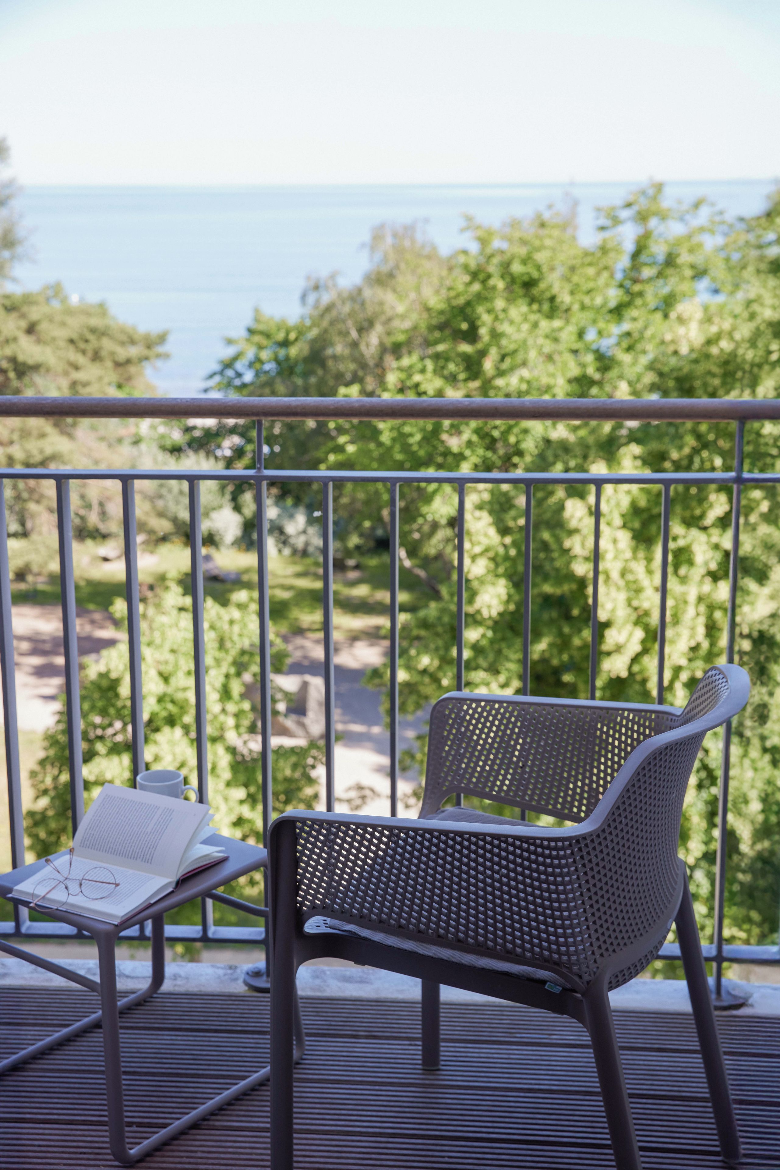 En balkon med to stole, hvoraf den ene har en bog og glas, med udsigt over træer og havet.