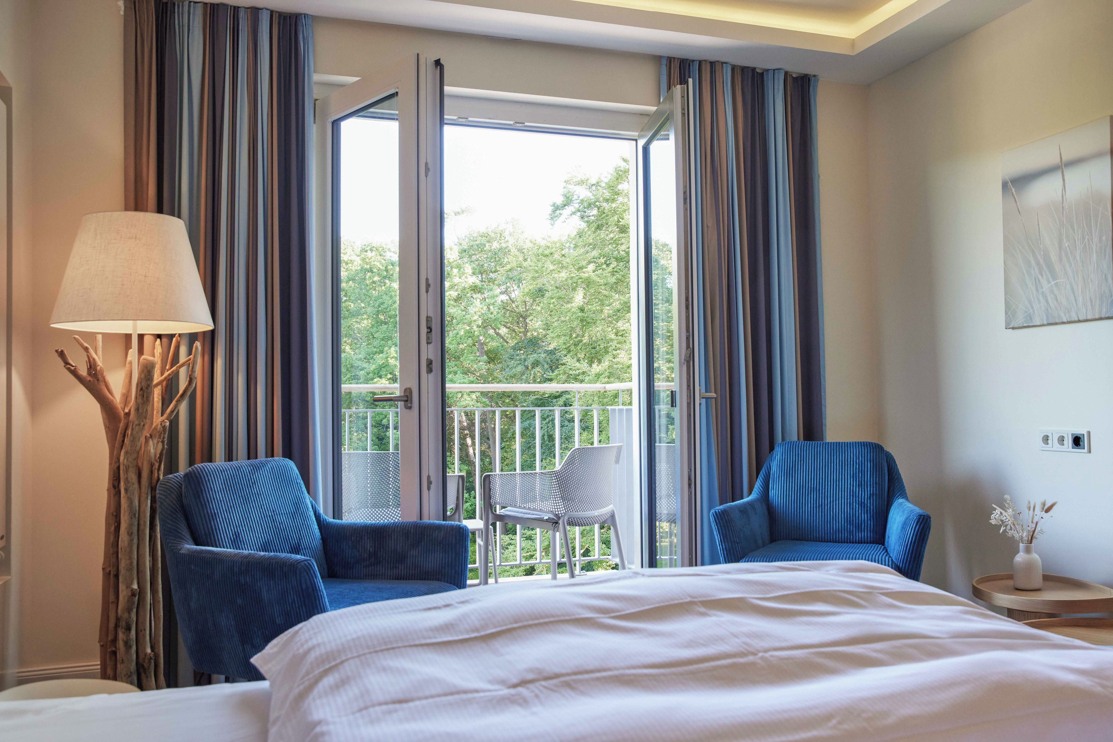 A hotel room with a bed, two chairs, a lamp, and a balcony with a view of trees.