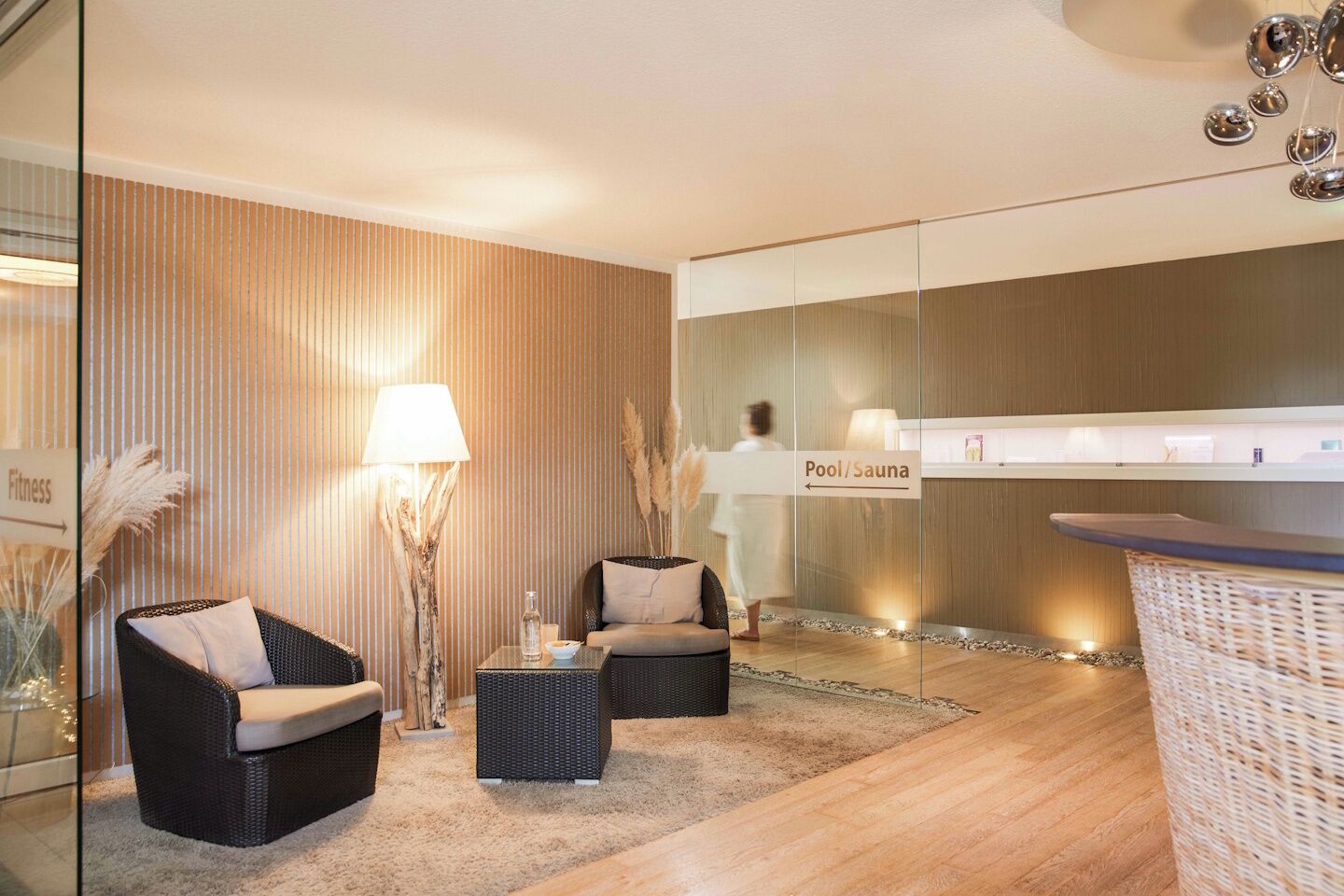 A modern hotel lobby with a reception desk, two wicker sofas, a lamp, and a sign indicating a pool/sauna area.