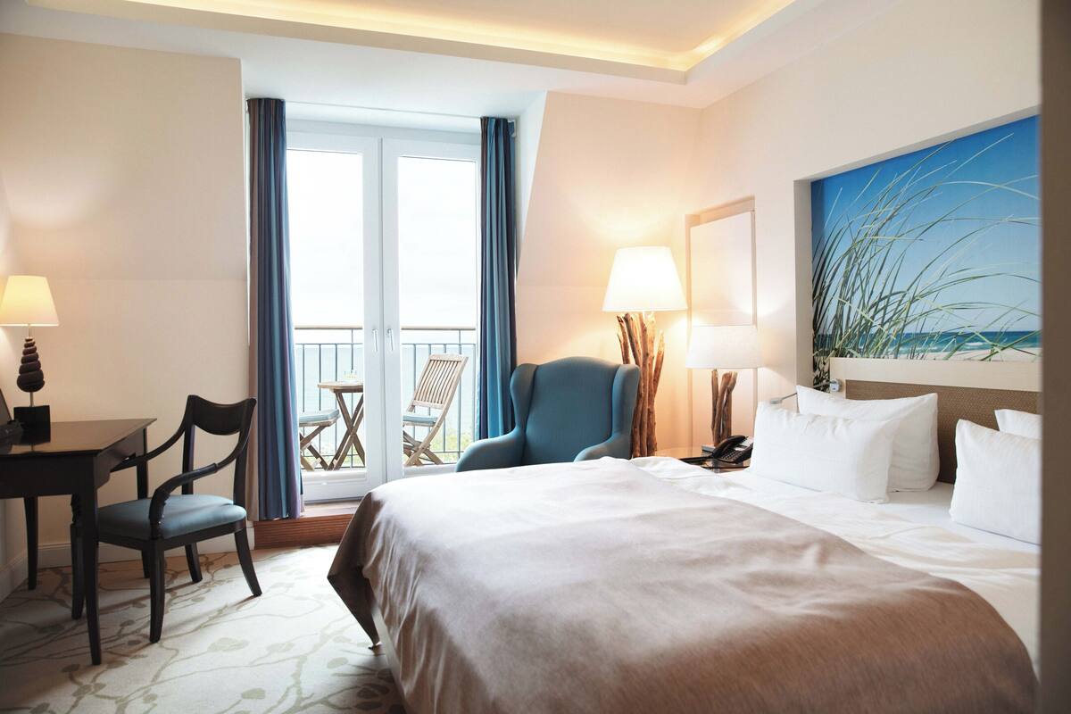 Une chambre d’hôtel avec un lit, un bureau et une chaise, un balcon avec vue et un tableau au mur.