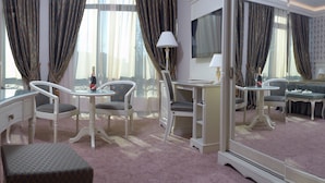 Habitación de hotel con un espejo grande, un televisor, un escritorio con una silla, una mesita redonda pequeña con una silla y una cama con cubrecama verde.