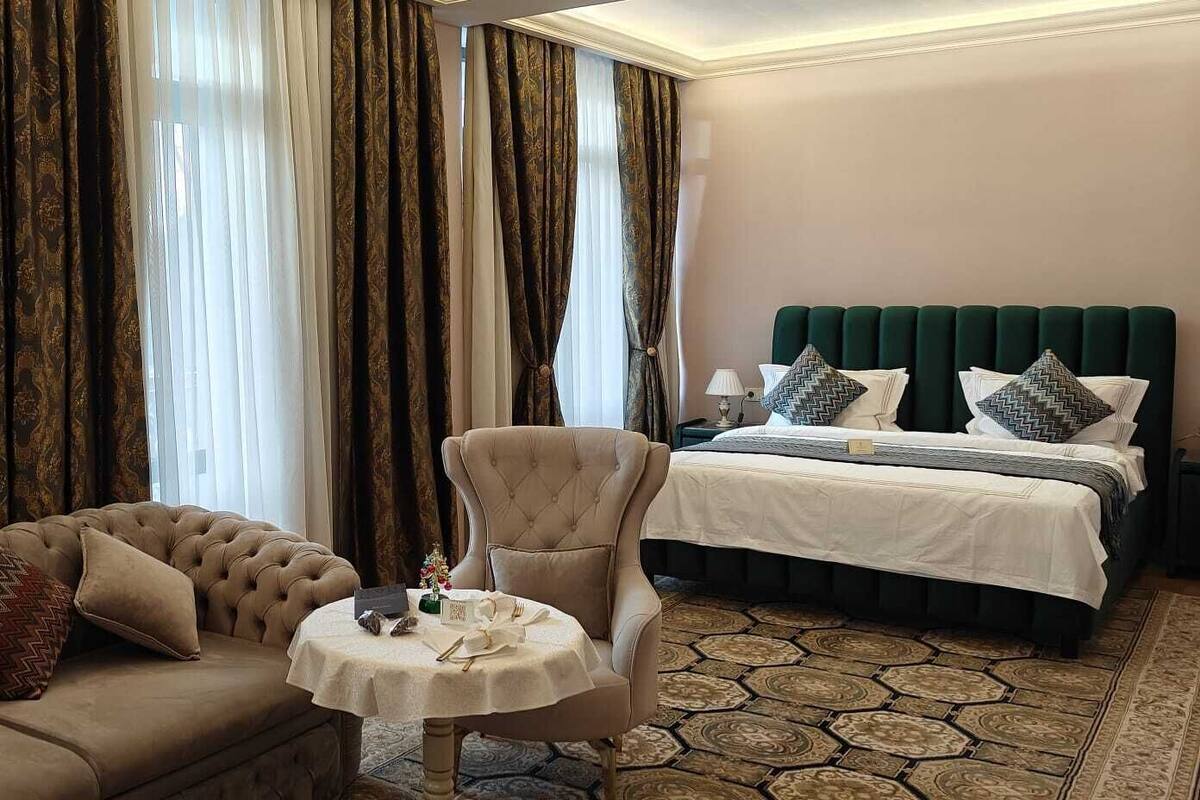 A hotel room with a tufted sofa, a round table with a vase, a green upholstered headboard, and a patterned rug.