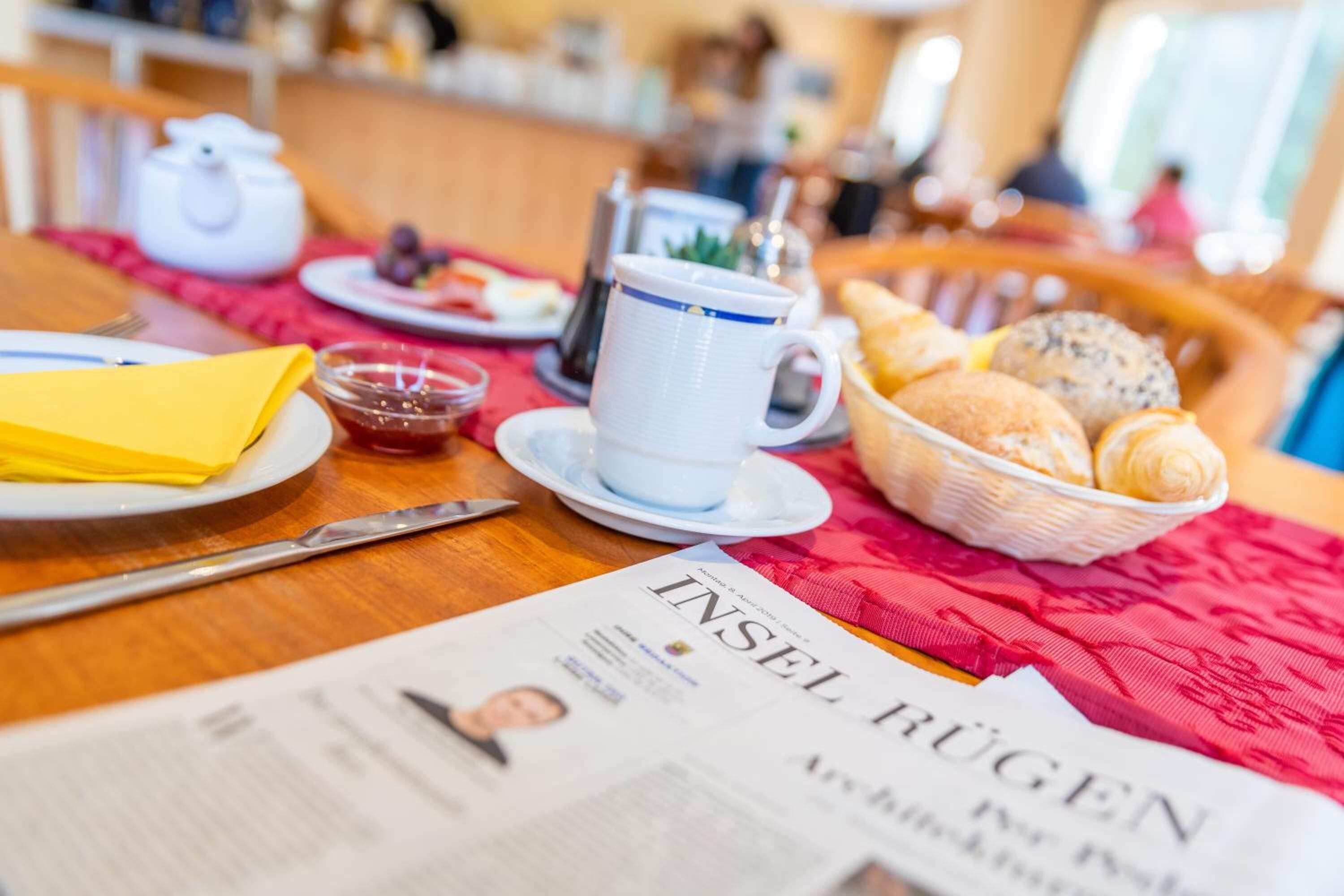 A breakfast table with a newspaper, a teapot, a cup, a basket of pastries, and a plate with a yellow napkin.