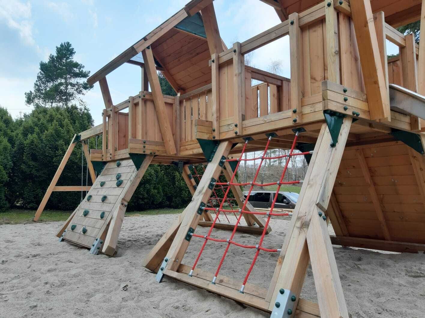 A wooden play structure with climbing nets and slides, set on sandy ground with trees in the background.