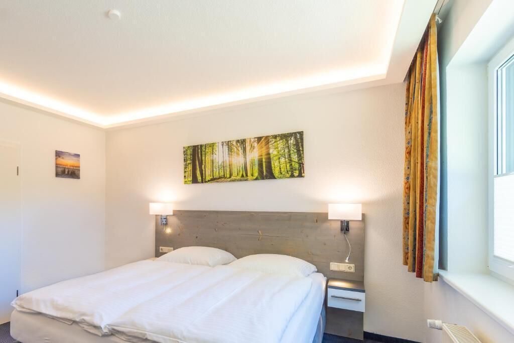 A hotel room with a neatly made bed, a headboard with wall-mounted lamps, a painting of a forest, and a window with curtains.