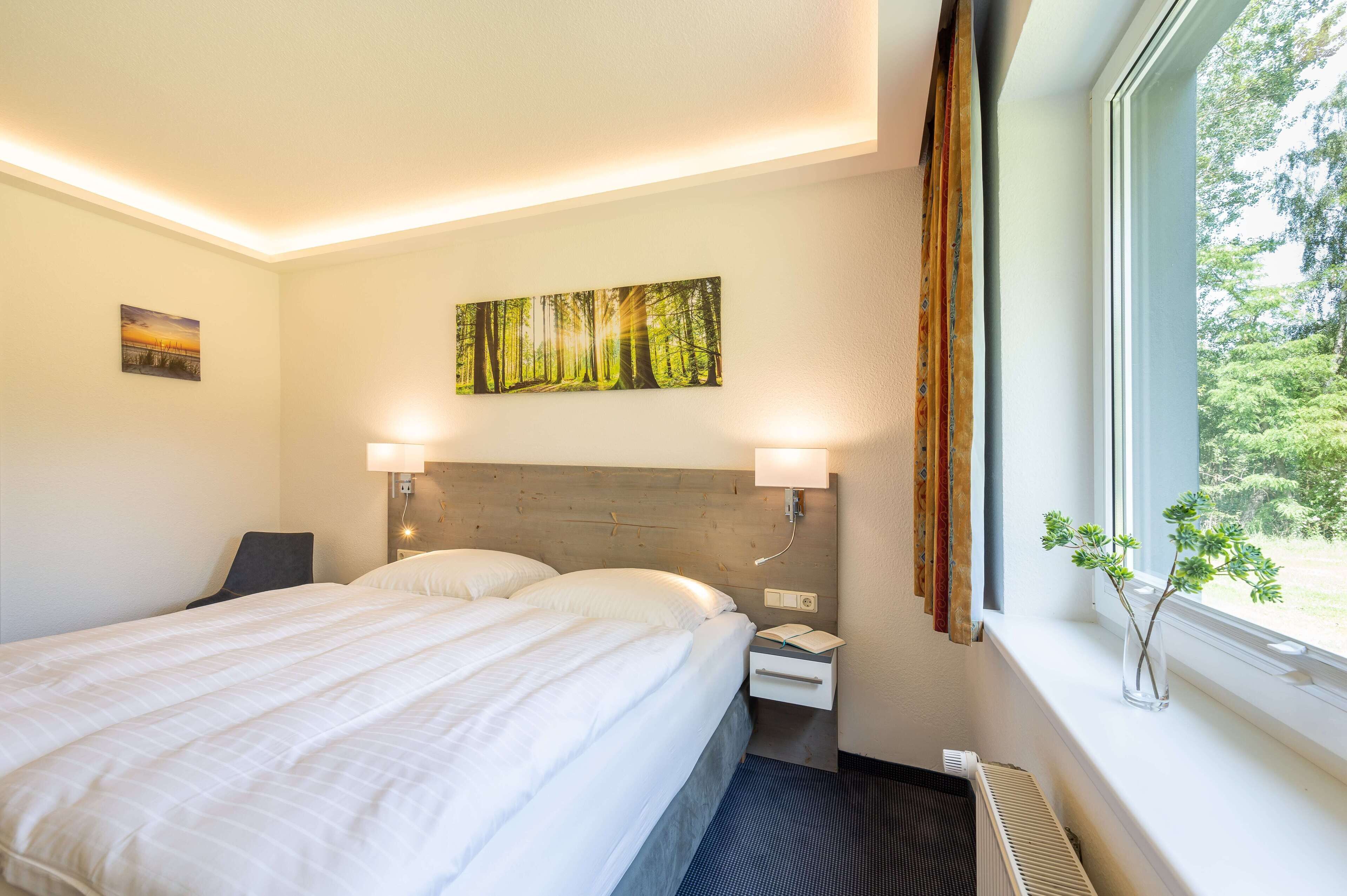 Een hotelkamer met een bed, nachtkastje, lamp en een boslandschap op het doek.