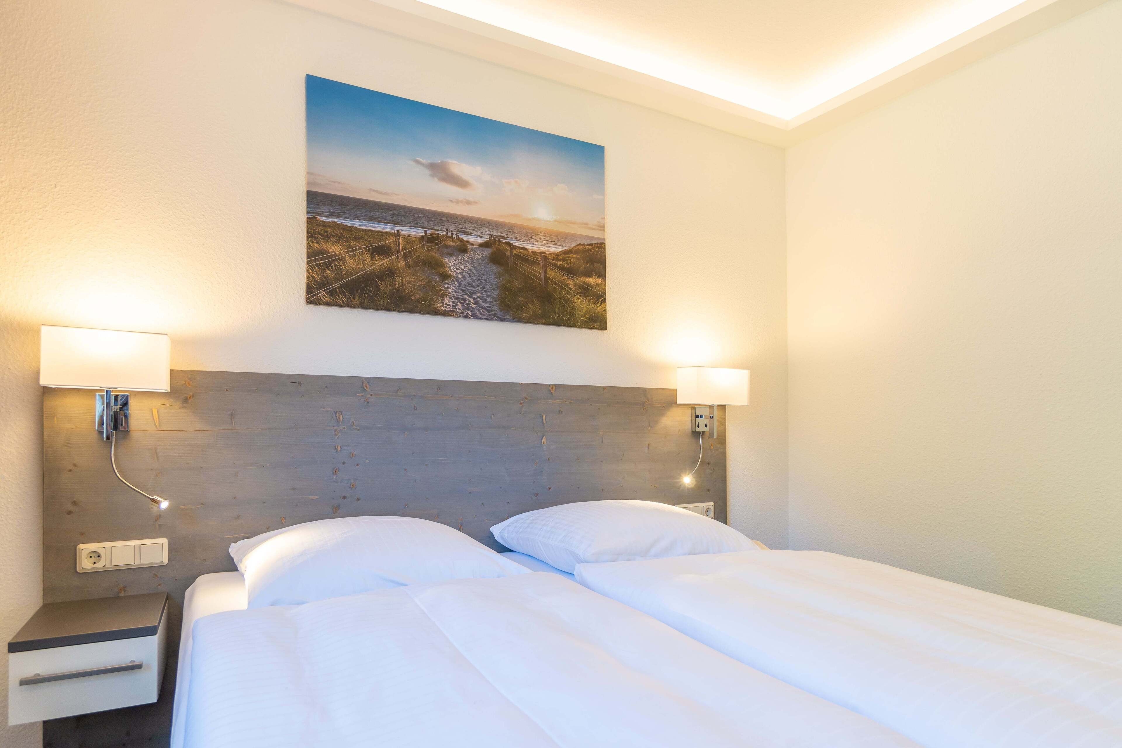 Habitación de hotel con cabecera de madera, dos lámparas de noche, una mesita de noche con cajón y un cuadro con una escena de playa sobre la cama.