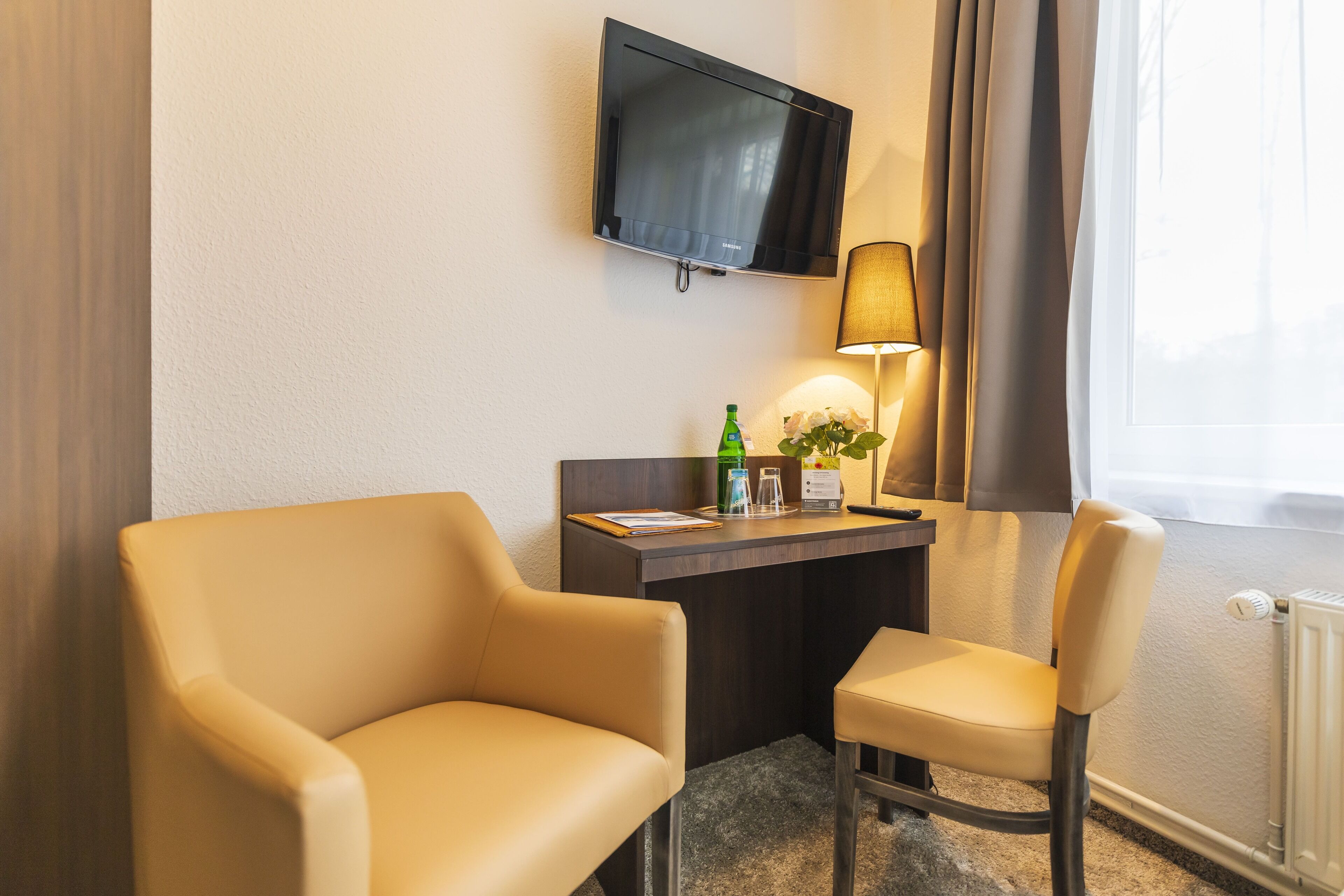 A hotel room with a desk, two chairs, a television, a lamp, and a window with curtains.