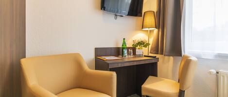 A hotel room with a desk, two chairs, a television, a lamp, and a window with curtains.