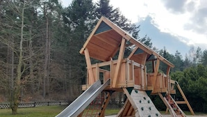 A wooden play structure with a slide and climbing areas, surrounded by trees and a fenced area. - Hotel Mukran (Sassnitz)