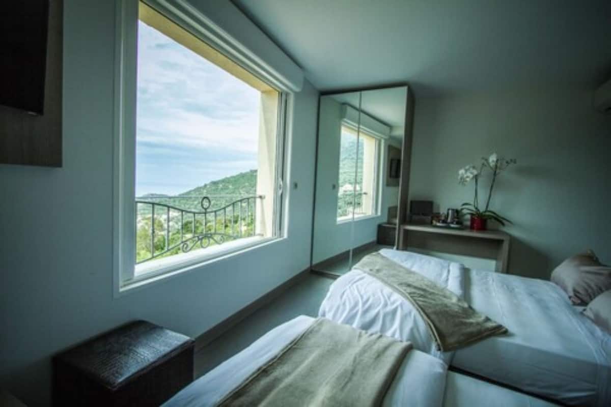 A bedroom with a large window, a bed, a bedside table, a mirror, and a view of greenery.