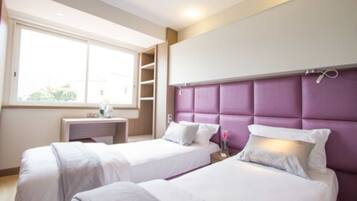 Habitación de hotel con dos camas, una ventana, una mesita de noche y un cabecero morado.