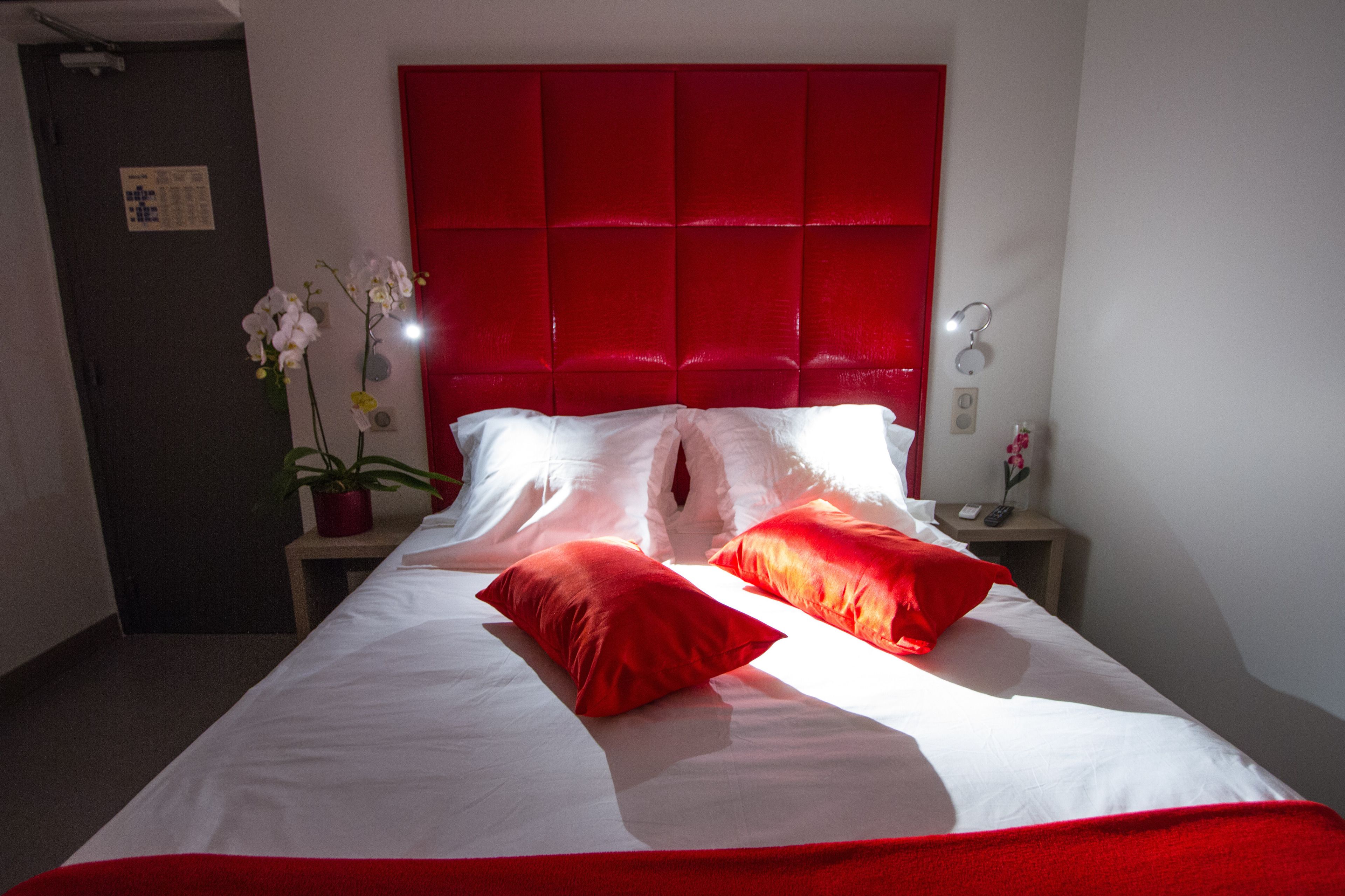 A hotel room with a red headboard, white bedding, and a small bedside table with a plant and a phone.