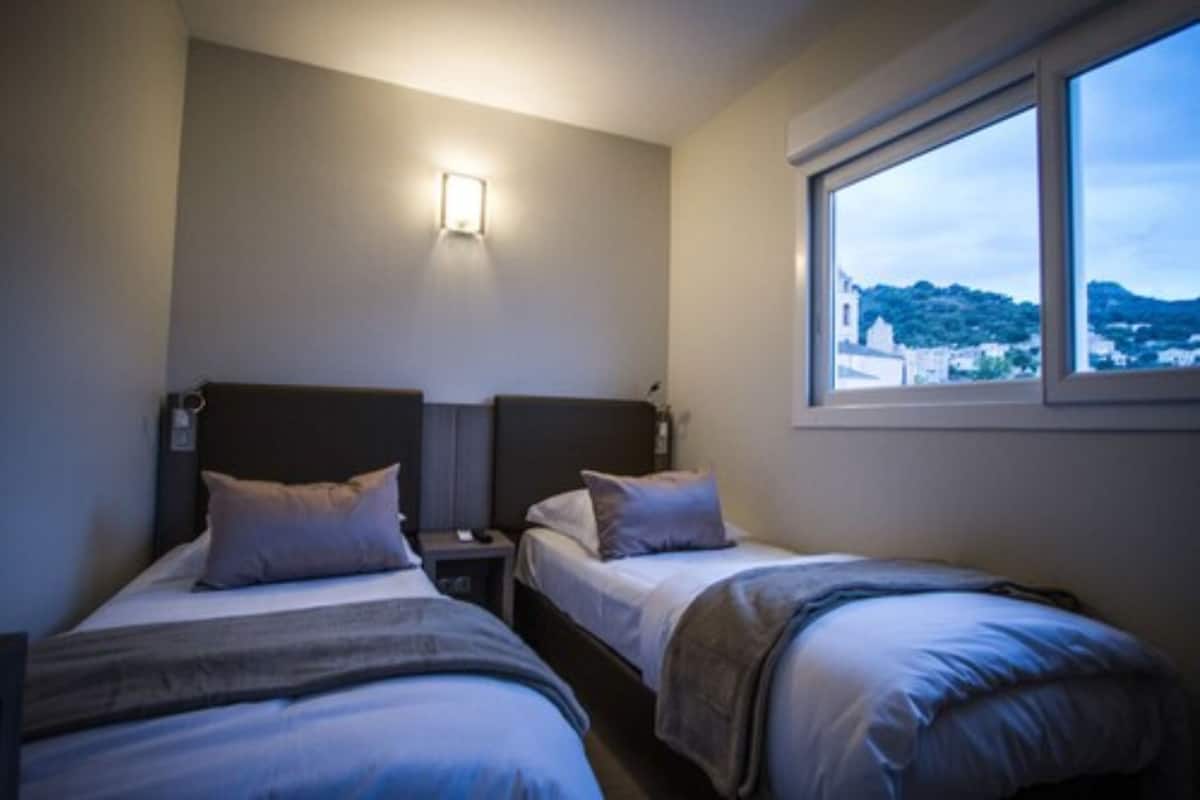 A hotel room with two beds, a nightstand, a wall-mounted light, and a window with a view of a town.