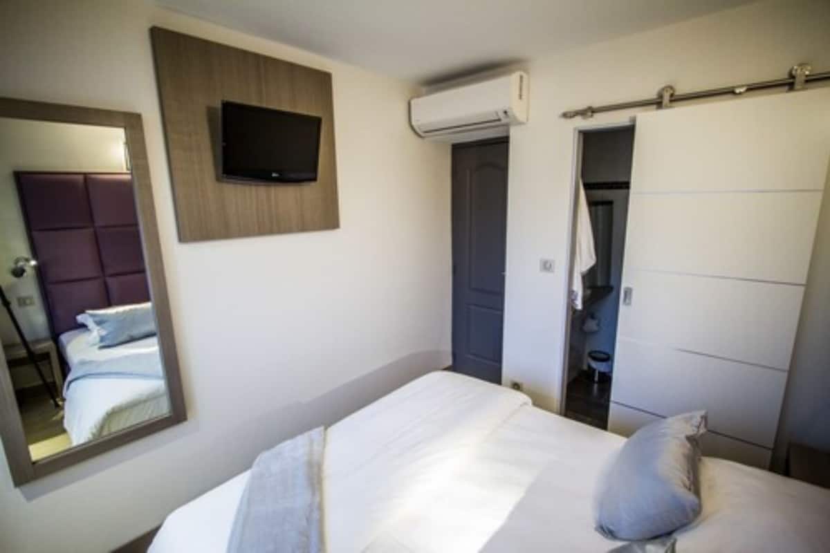 A hotel room with a bed, a television mounted on the wall, an air conditioning unit, and a sliding door leading to a bathroom.