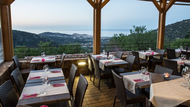 Un espace de restauration en plein air avec des tables pour deux, offrant une vue panoramique sur les collines et la mer.