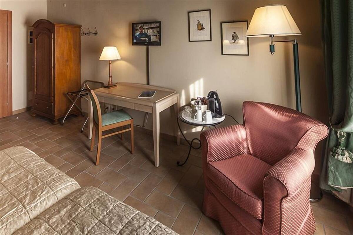 A hotel room with a bed, a desk with a chair, a red armchair, a lamp, a small table with a kettle, and a wardrobe.
