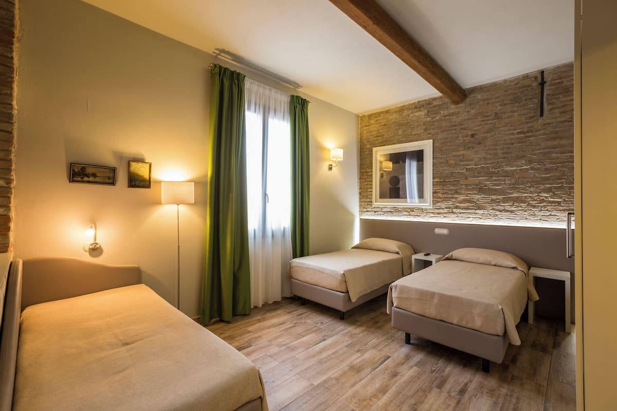 A room with two single beds, a large window with green curtains, a stone wall, and a small bedside table with a lamp.
