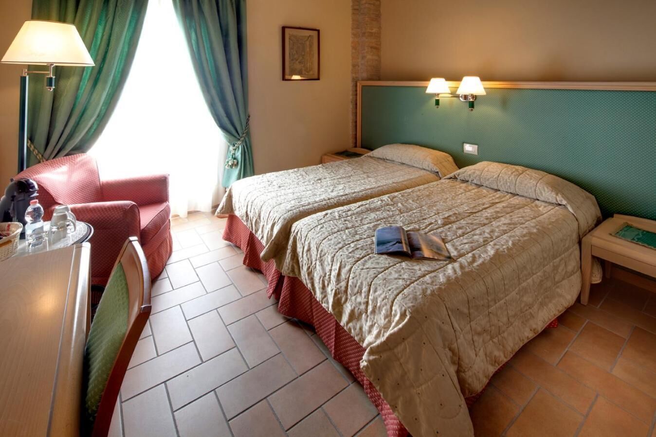 Habitación de hotel con dos camas, un escritorio con lámpara, una silla y una ventana con cortinas.