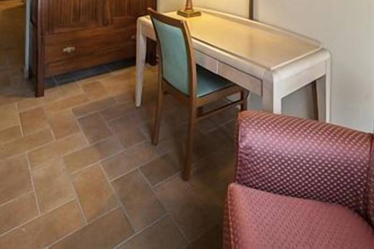 A hotel room with a wooden wardrobe, a desk with a lamp, a television mounted on the wall, and a patterned chair.