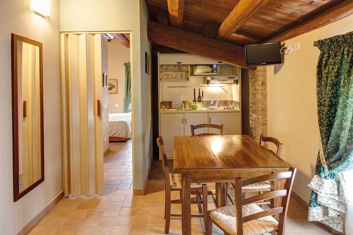 A dining area with a wooden table and chairs, a kitchen in the background, and a television mounted on the wall.