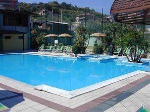 A swimming pool with lounge chairs and umbrellas, surrounded by greenery and buildings. - Villaggio Verde Cupra (Cupra Marittima)